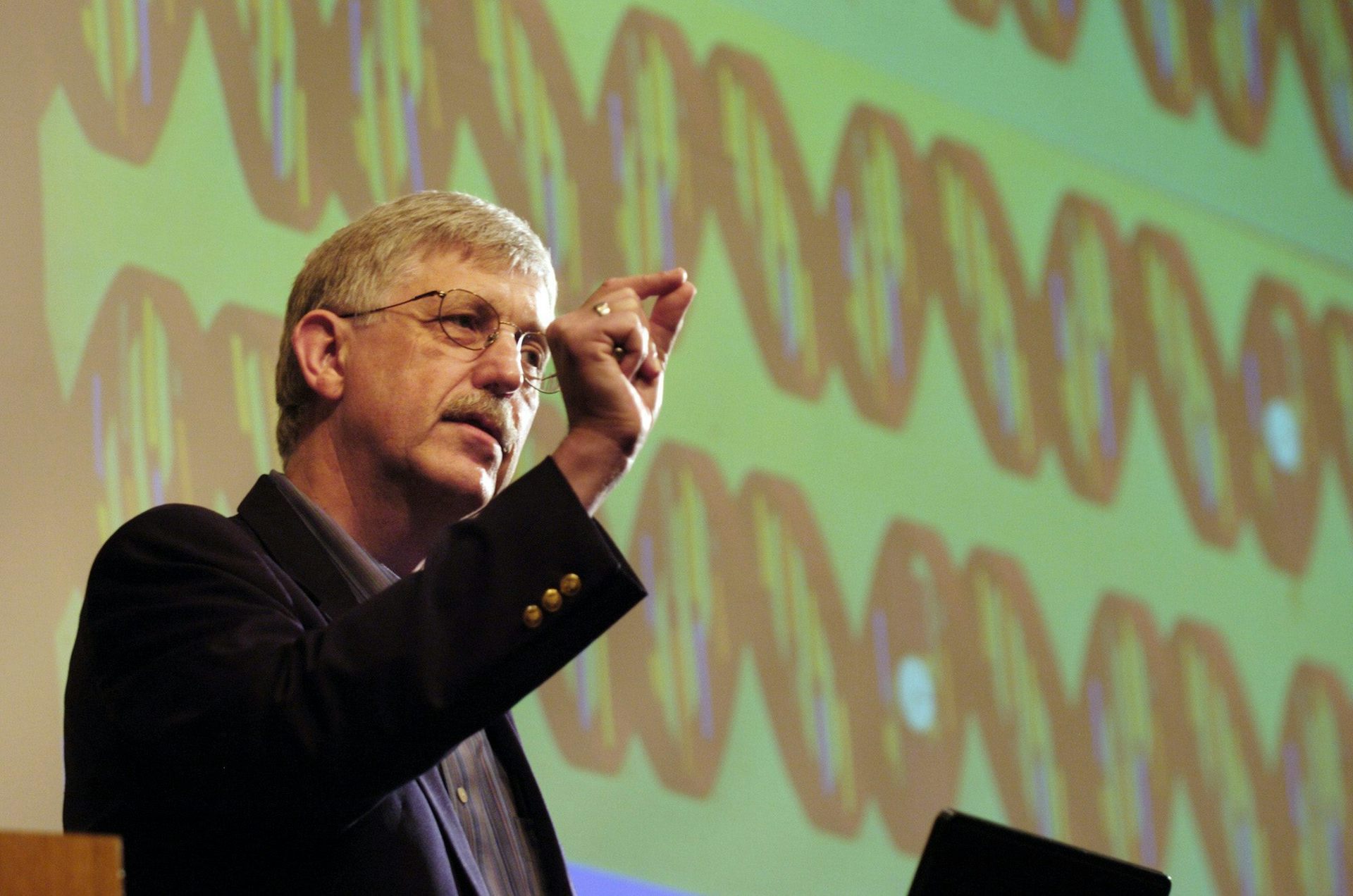 Un hombre caucásico con bigote y gafas da un discurso mientras está de pie frente a una presentación que muestra los cromosomas.