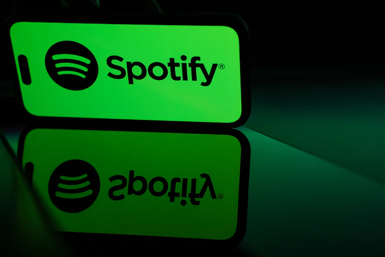 A mobile phone with a green Spotify logo on the screen is reflected off a shiny surface.