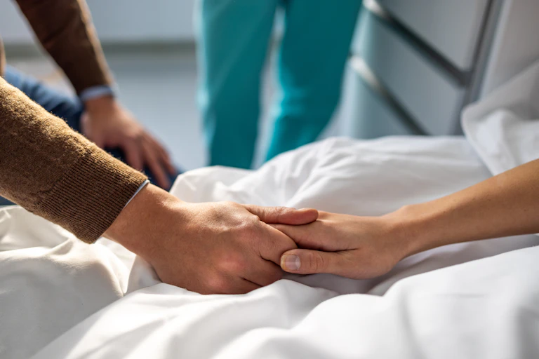 Someone in a brown sweater holds the hand of someone lying in a hospital bed.