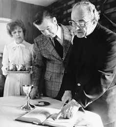 A black and white photograph shows a middle-aged woman in a white dress and a middle-aged man in a suit jacket, speaking with a man in a black clerical uniform.