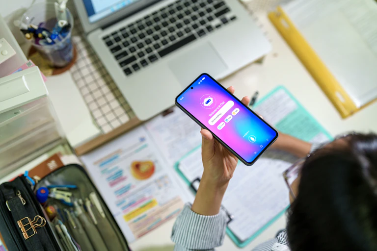 A student at a desk with laptop and writing materials ask a chatbot a question on a phone.