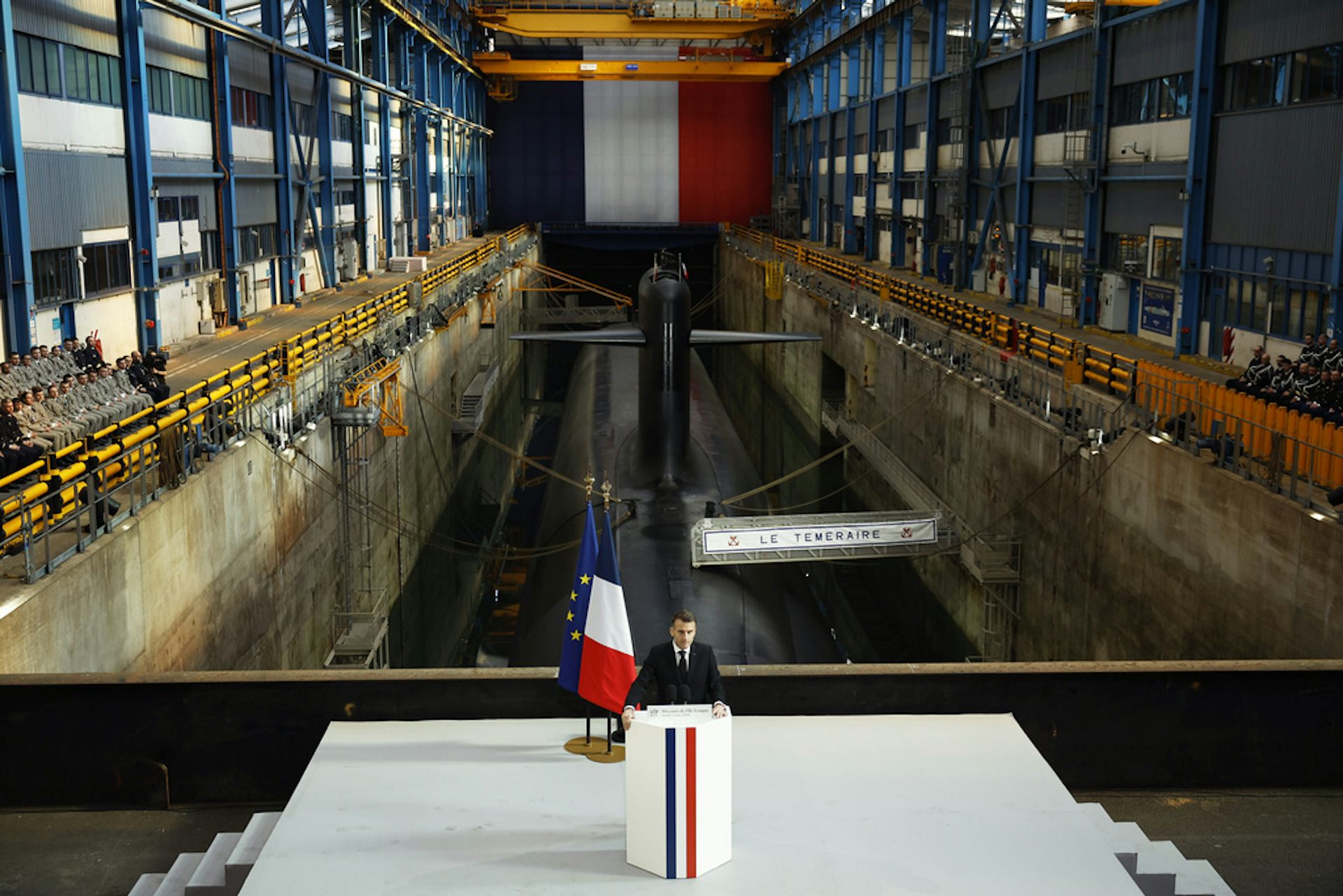 A suited man gives a speech at a lectern in front of a submarine.