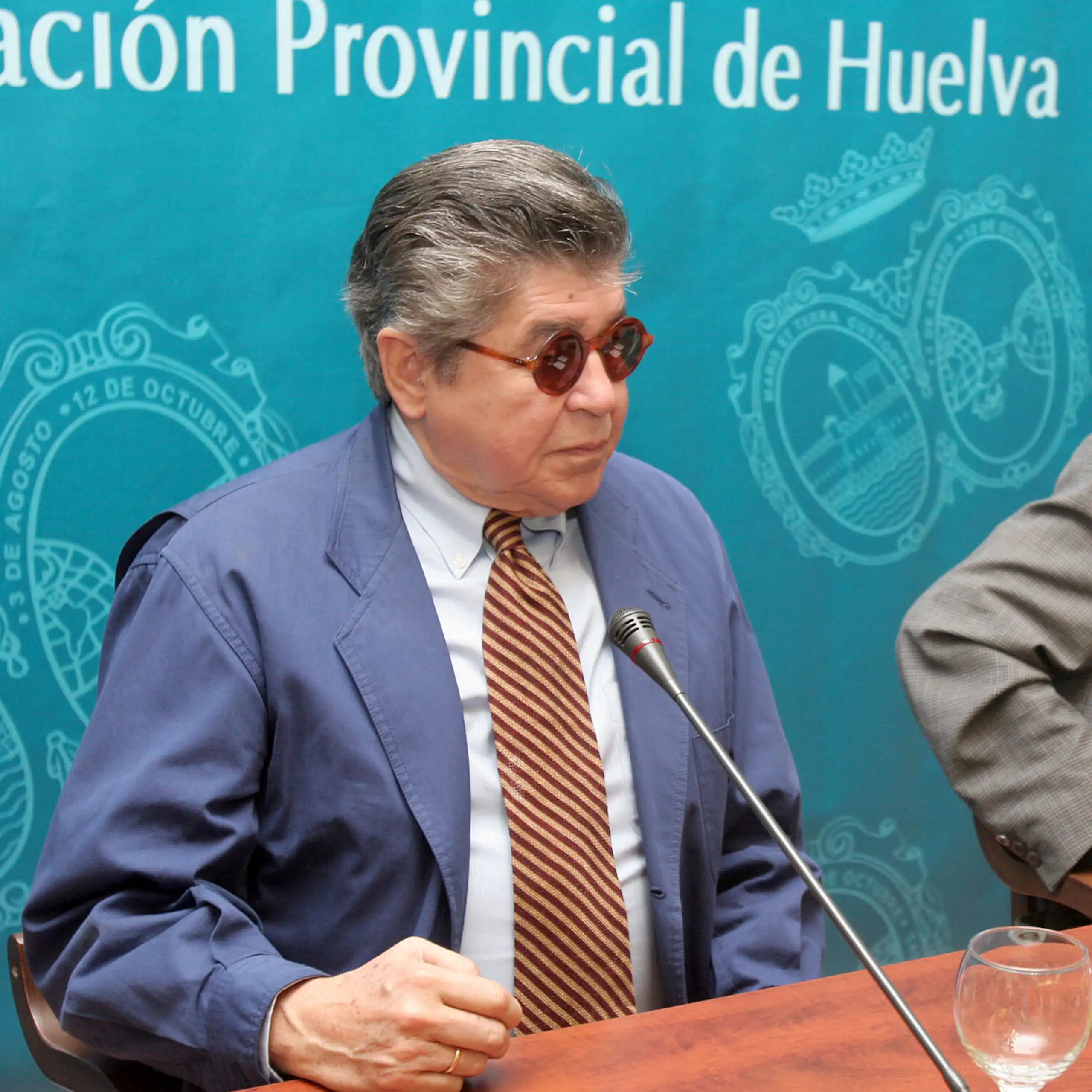 Un hombre con traje y gafas de sol a punto de hablar delante de un micro.