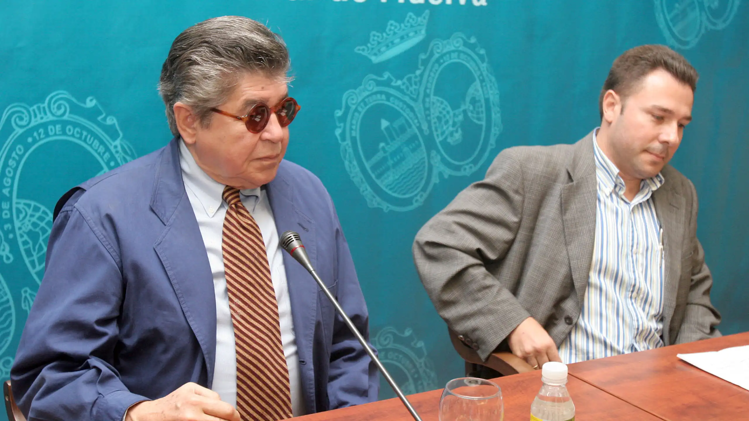Un hombre con traje y gafas de sol a punto de hablar delante de un micro.