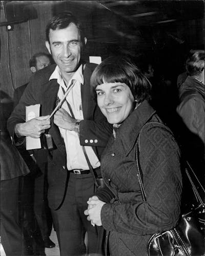 A man and woman in an airport, carrying bags and papers.