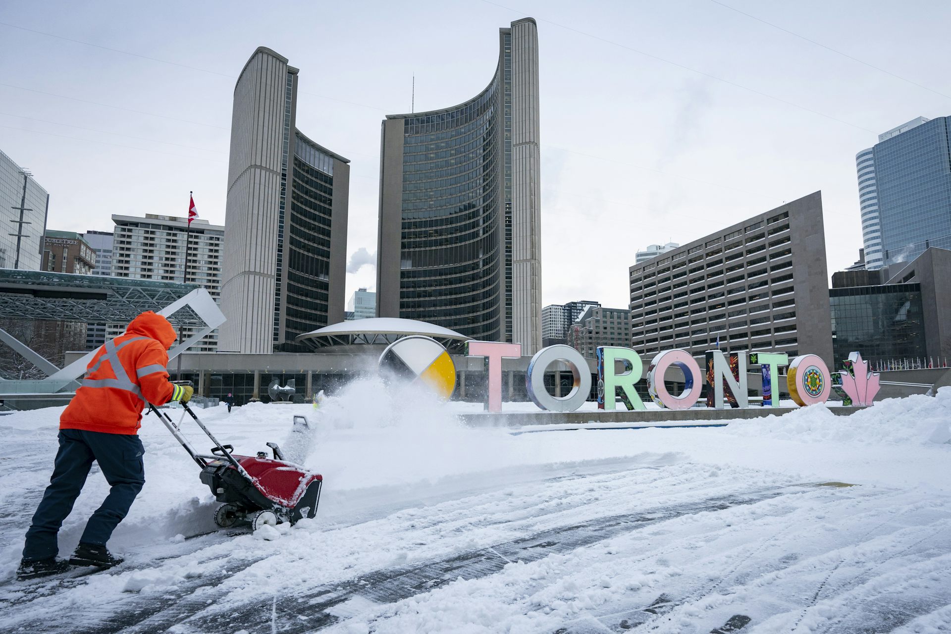 A city worker in an orange vest uses a snowblower in front of Toronto city hall.