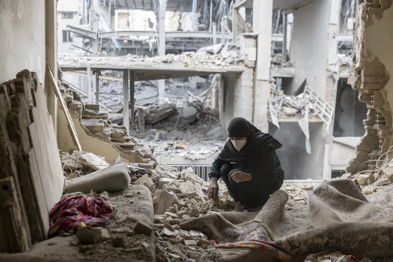 A woman crouches down picking up up pieces while surrounded by missing walls and pulverized cement. Few items int he home are reconizable.