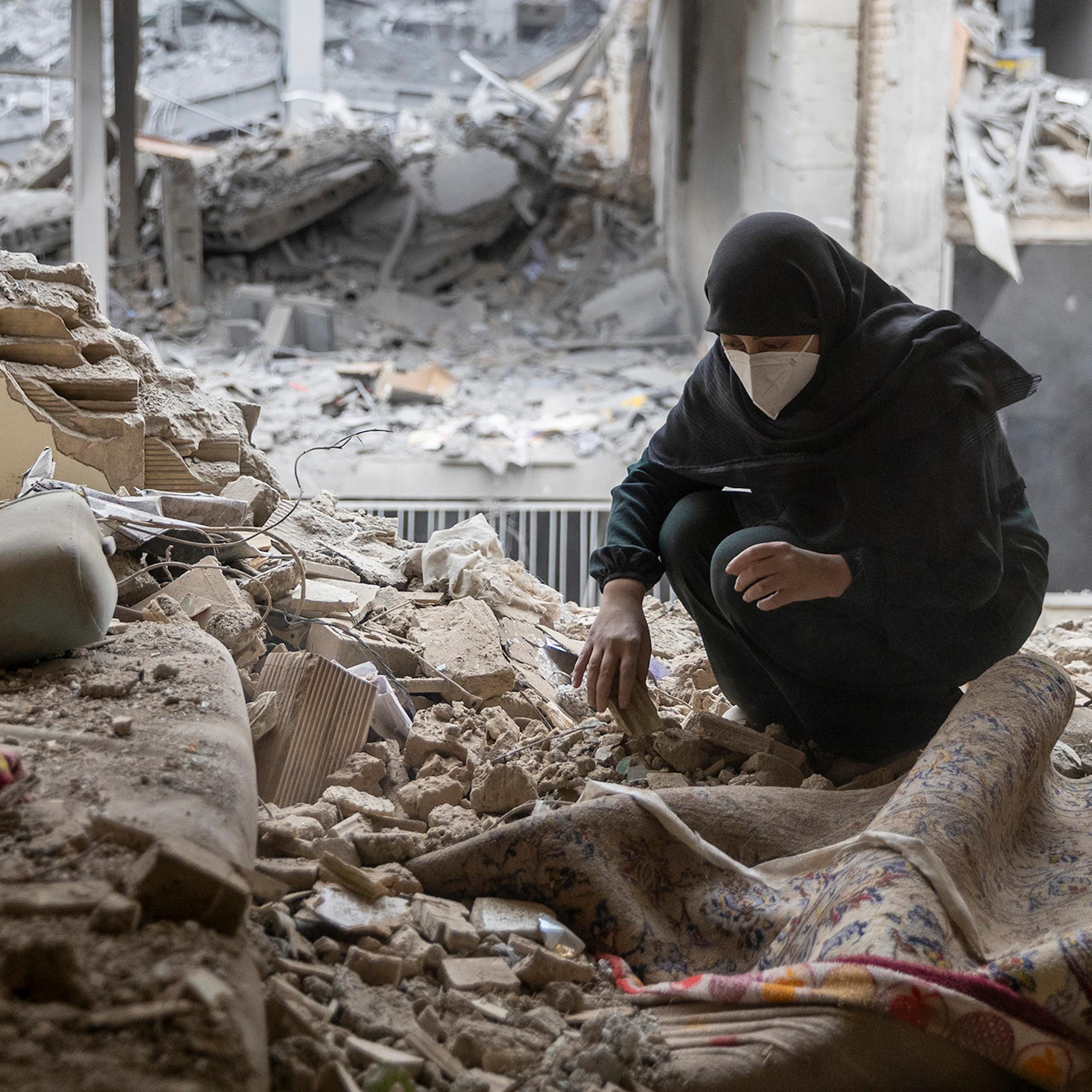 A woman crouches down picking up up pieces while surrounded by missing walls and pulverized cement. Few items int he home are reconizable.