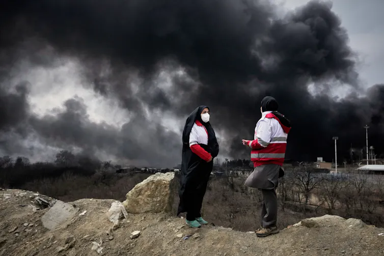 Two women in hijabs and white/red jackets stand with a large plume of black smoke behind them
