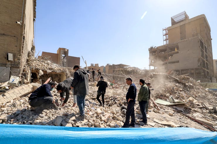 A group of men walk through the remains of a building that collapsed. Several buildings around them are also damaged.