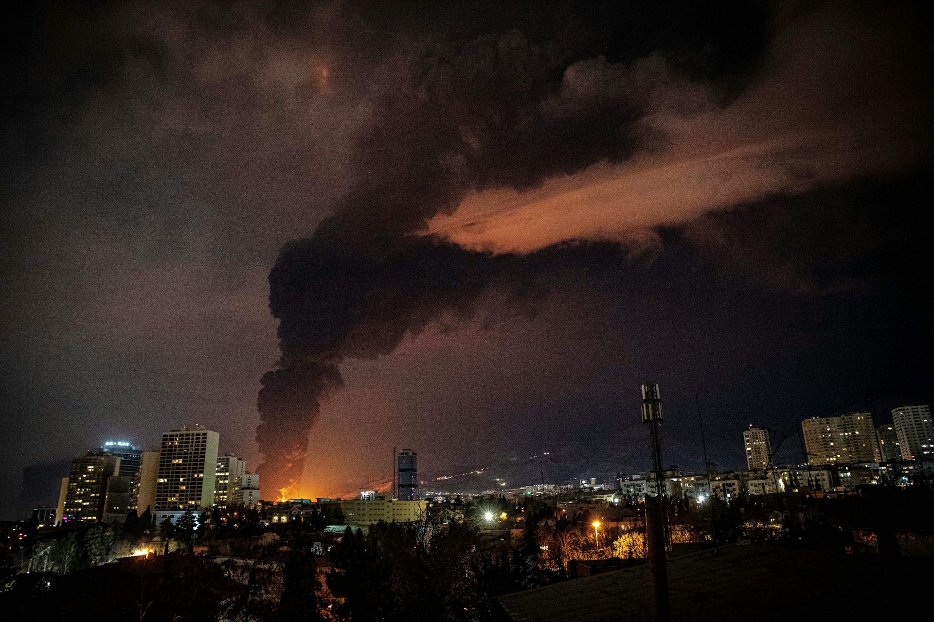 A large plume of black smoke rises above a city at night