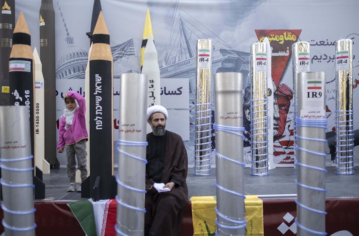 A young girl and a man in a black robe and white turban stand next to tubes decorated to look like missiles and centrifuges.