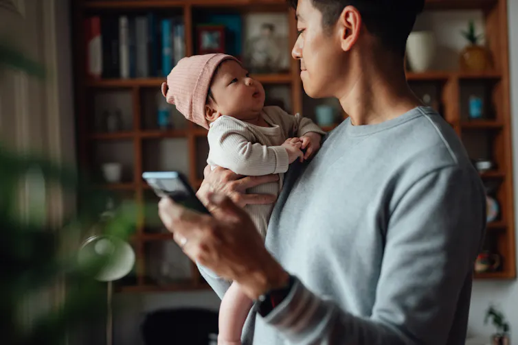 An Asian man in a gray sweatshirt smiles at a baby as he holds a phone.
