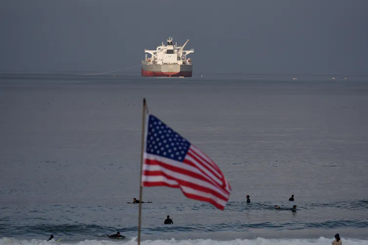 Una bandera estadounidense ondea en primer plano mientras un petrolero navega al fondo.