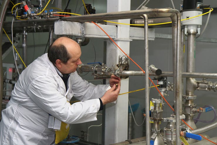 A person in a white coat puts his hands on some metal piping.