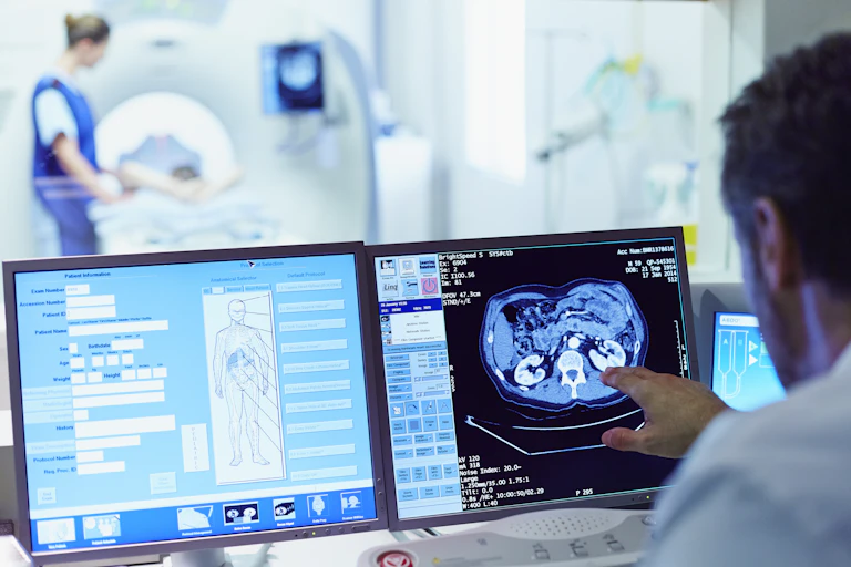 Doctor running CT scan from the next room, with patient entering a CT scanner in the background.