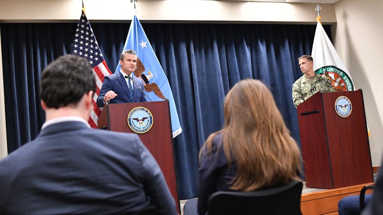 Two men, including one in a military uniform, at lecterns, speaking.