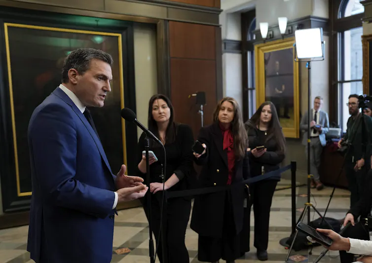 Un hombre con traje azul está detrás de un micrófono con otras personas al fondo.