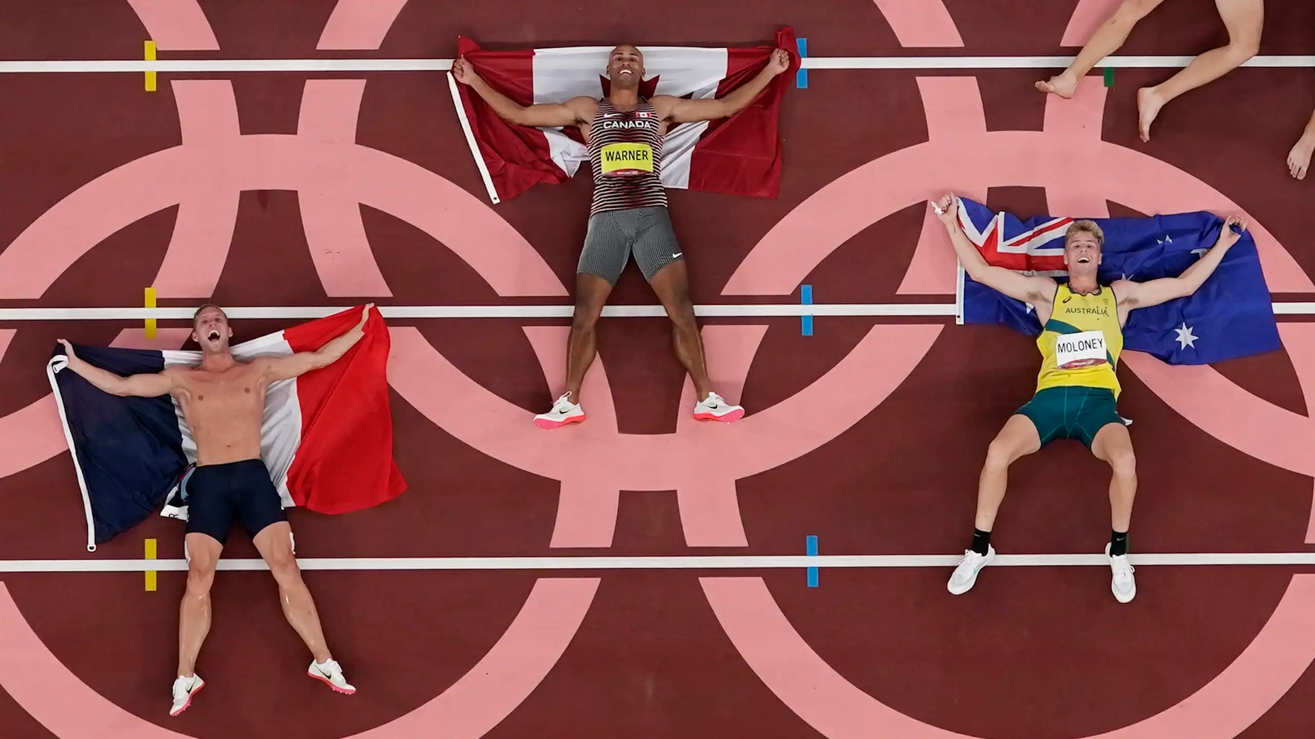 Three men with flags lying on a track inside the circles of the Olympic rings