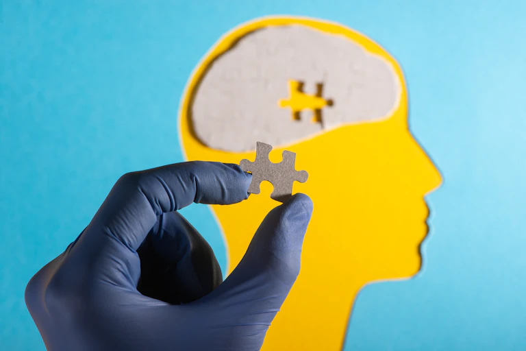 A cutout of a head in side-profile. In the centre of the head's brain, it is missing a puzzle piece. A person's hand holds that missing piece between their thumb and forefinger.