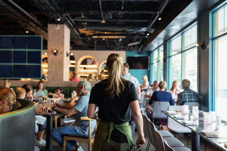 A waitress in a busy restaurant, ponytailed back to camera