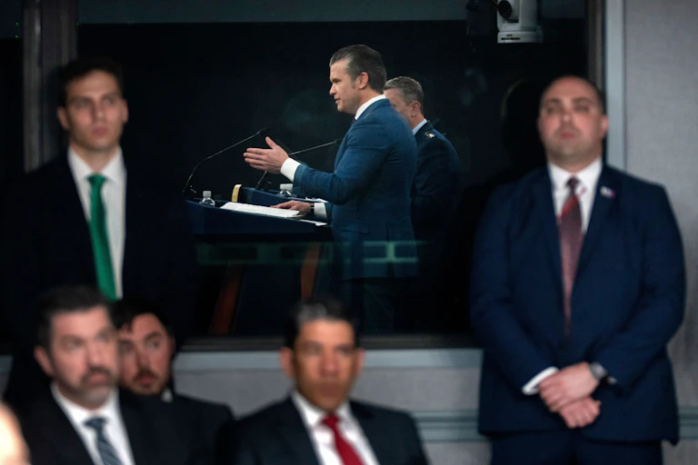 Several men in suit and ties watch another man speak while on a podium.