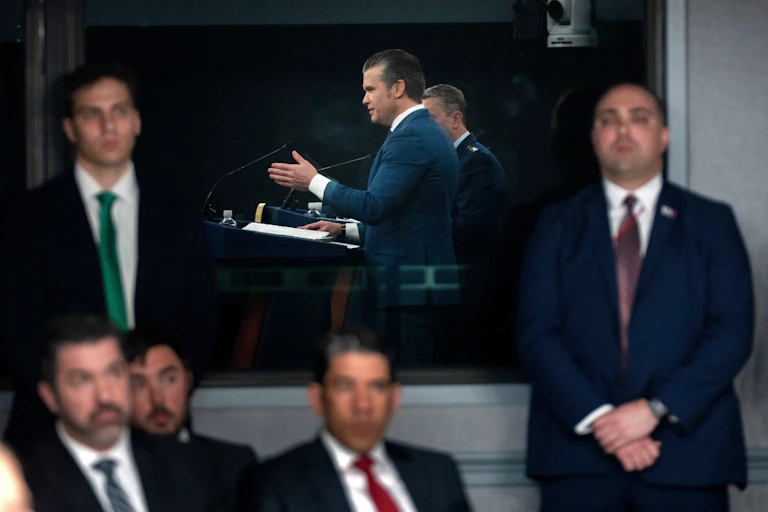 Several men in suit and ties watch another man speak while on a podium.