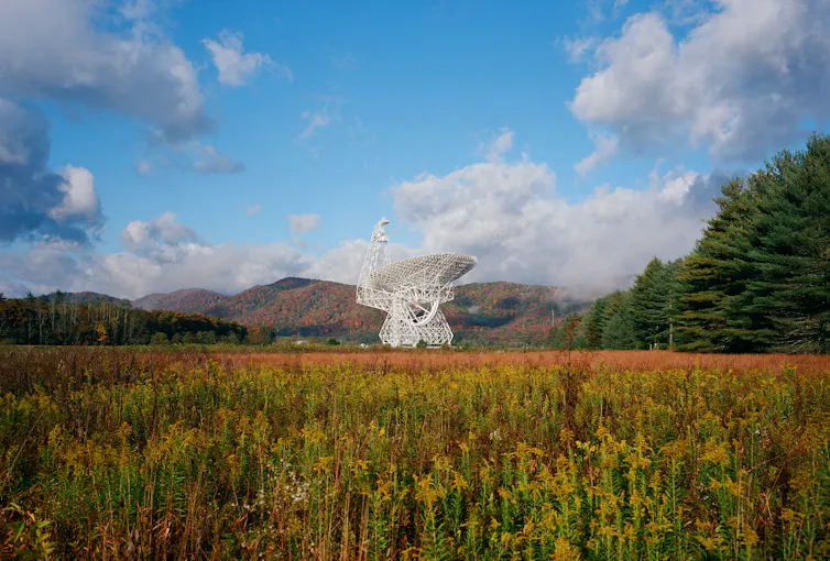 Large white radio telescope at center with cloudy blue sky overhead and orange, green, and yellow field in the foreground. Mountains are in the background, and a crop of trees are to the right of the image.