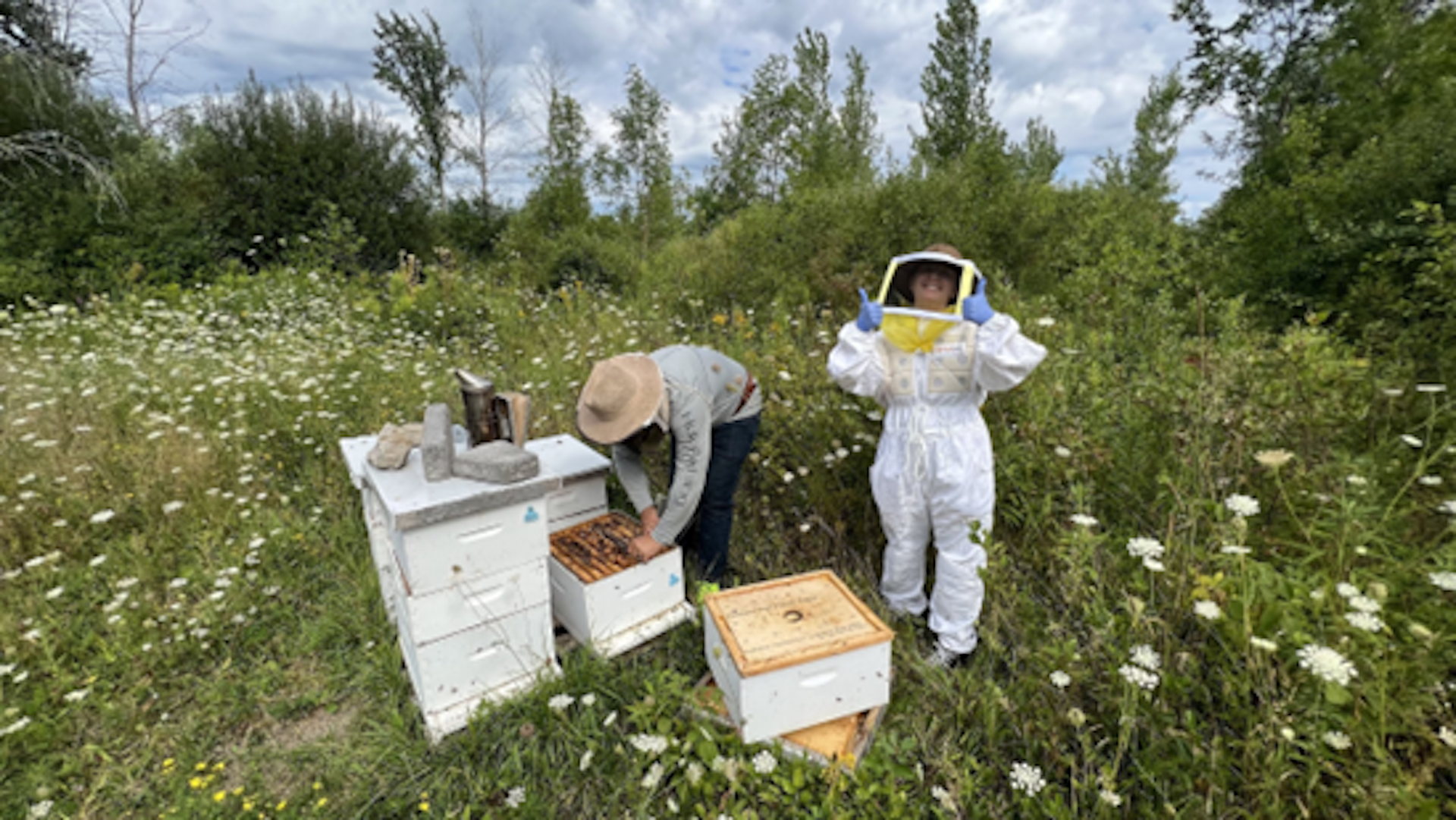 Brendan Daisley se inclina sobre una colmena abierta, Liz Mallory permanece parada con un traje de apicultura completo y con los pulgares hacia arriba.