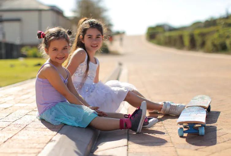 Two children in dresses sitting on curb on a neighborhood, skateboards before them