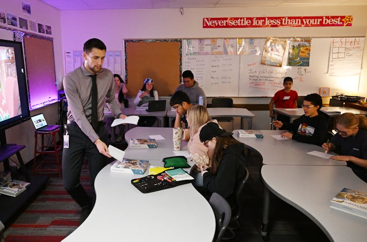 A man wearing a grey shirt and dark tie hands a piece of paper to teenagers seated at long white tables.