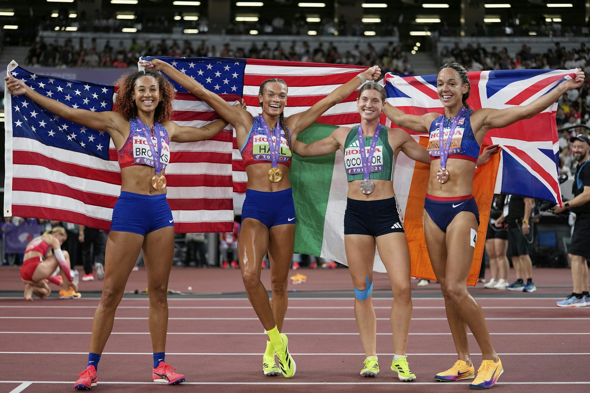 Cuatro mujeres vestidas con ropa deportiva, cubiertas con banderas en la pista de atletismo