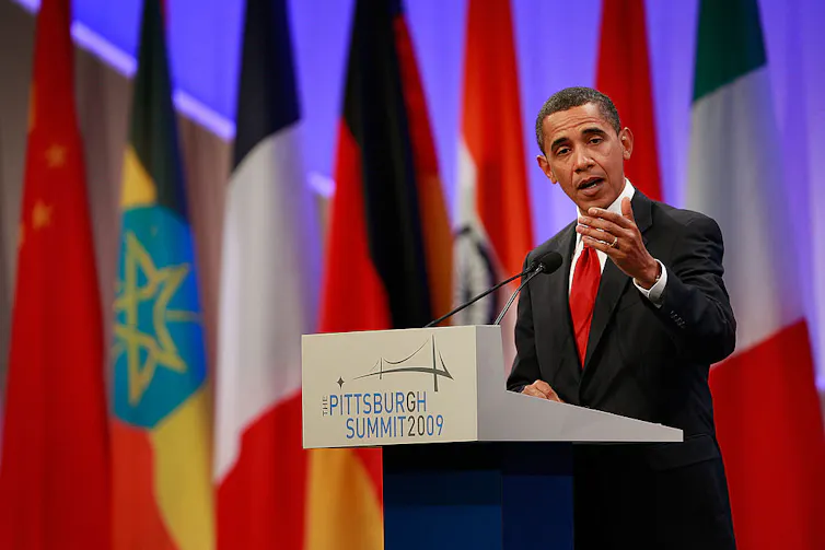 A thin Black man in a suit and red tie speaks at a podium with world flags behind him.