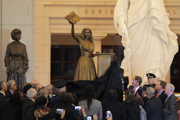 A statue of a teen girl holding up a book is unveiled in front of a lot of people.