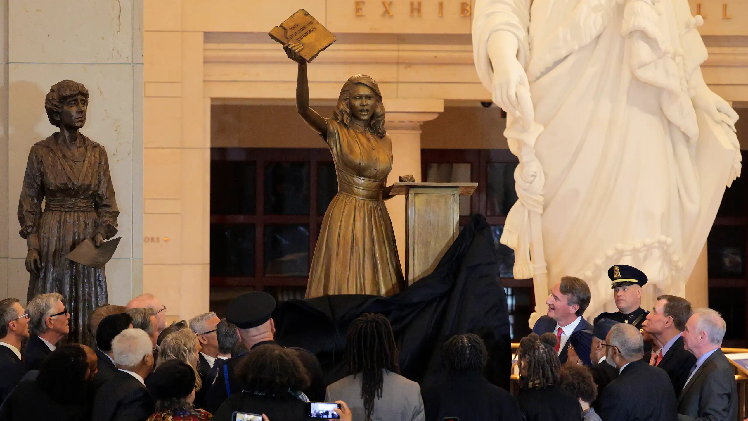 A statue of a teen girl holding up a book is unveiled in front of a lot of people.