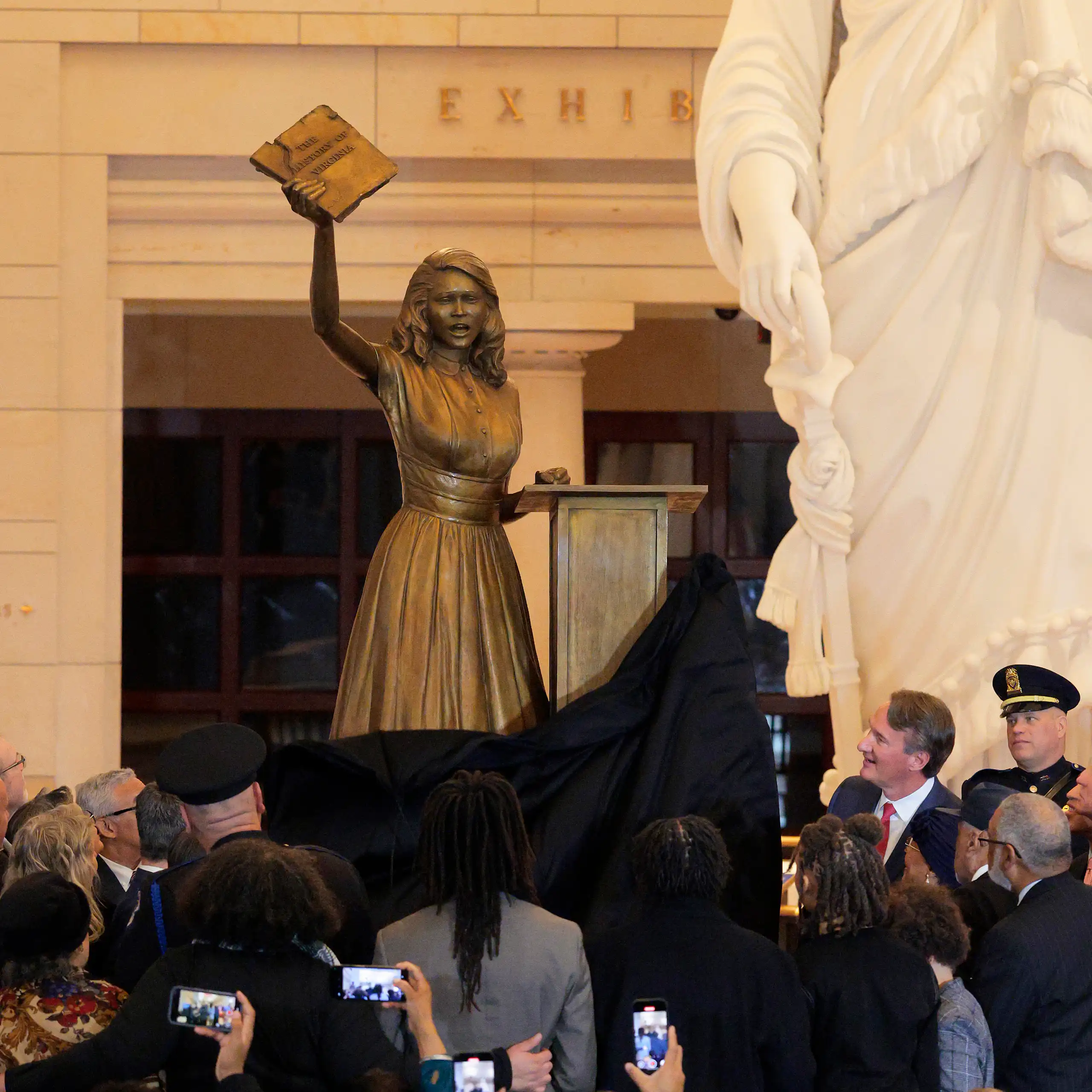 A statue of a teen girl holding up a book is unveiled in front of a lot of people.