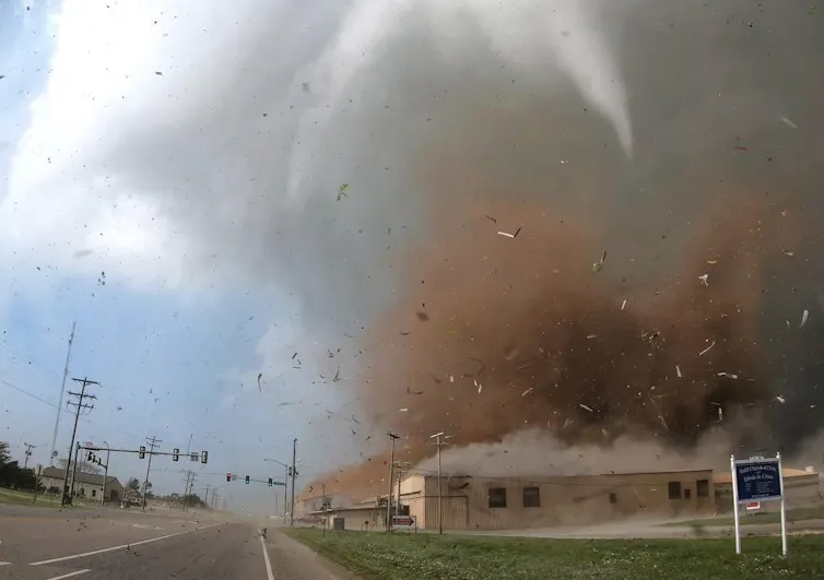 Um tornado destrói um prédio, lançando uma nuvem de detritos em alta velocidade no ar.