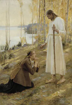 A man in a simple white robe, holding a walking stick, stands above a crouching women in brown garments by the shore of a lake.