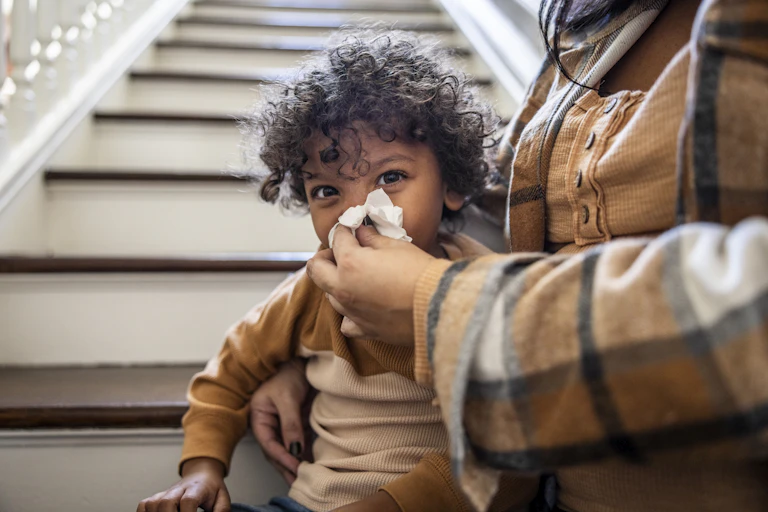 Woman's hand holding tissue to kid's face blowing nose.