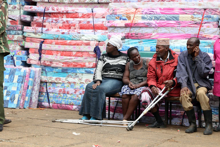 Four people, one of them with crutches, seated next to a pile of mattresses wrapped in plastic