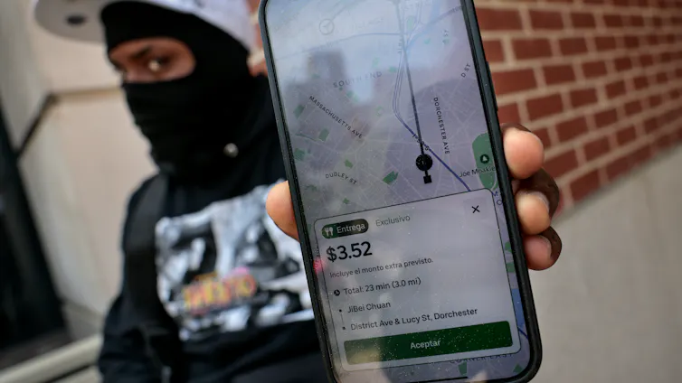 A food delivery gig worker holds up his smartphone with a food delivery order. The phone displays the information in Spanish.