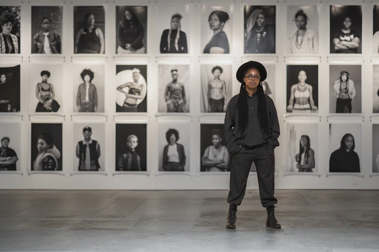 An African woman poses for a portrait against a wall of portraits in black and white.