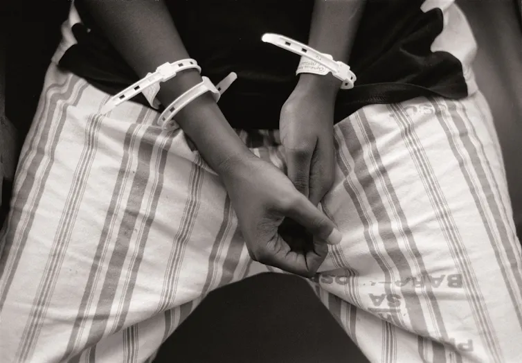 A black and white photo of a person holding their hands together, medical tags on their arms.