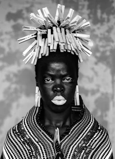 An African woman looks directly at camera, her hair a wig of pegs.