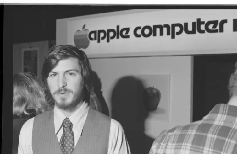 a young man with a beard and mustache wearing formal clothes stands in front of sign saying 'apple computer'