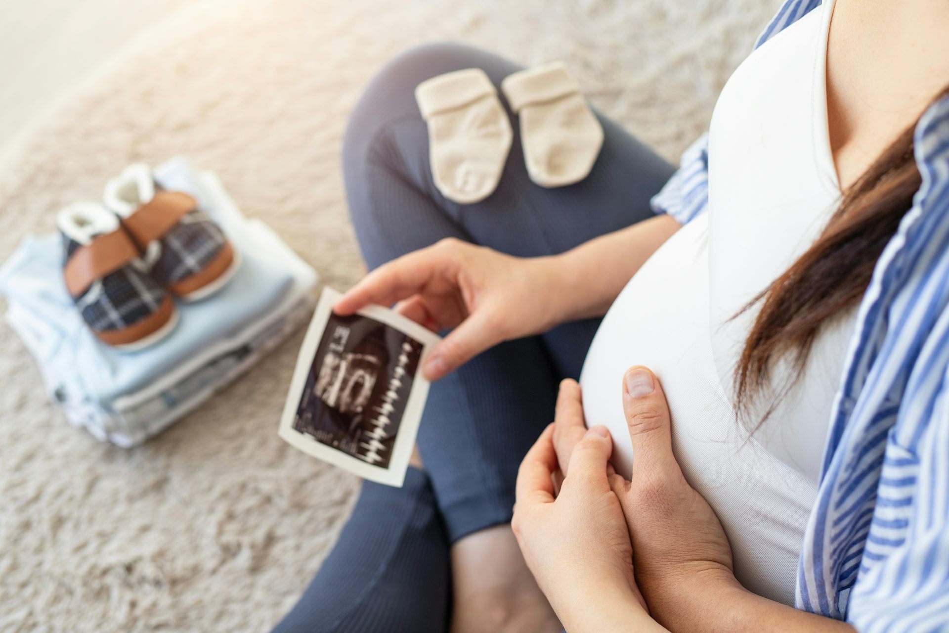 Une femme enceinte regarde la photo de l’échographie de son bébé, à côté d’elle, on voit des chaussettes et chaussons pour le bébé à naître.