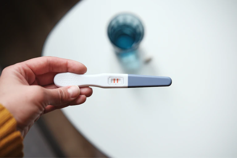 A person's hand holding up a positive pregnancy test with a glass of water and a pill sitting in the background.
