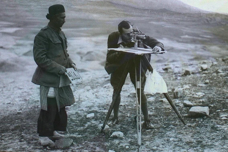 A black and white photo of a man using a telescope to survey an area while another stands beside him