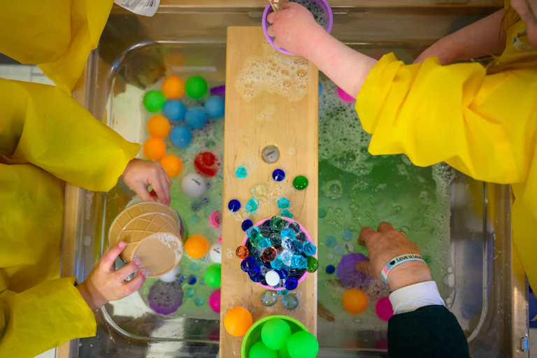 Young people's hands are seen in a bin of soapy water with plastic toys.