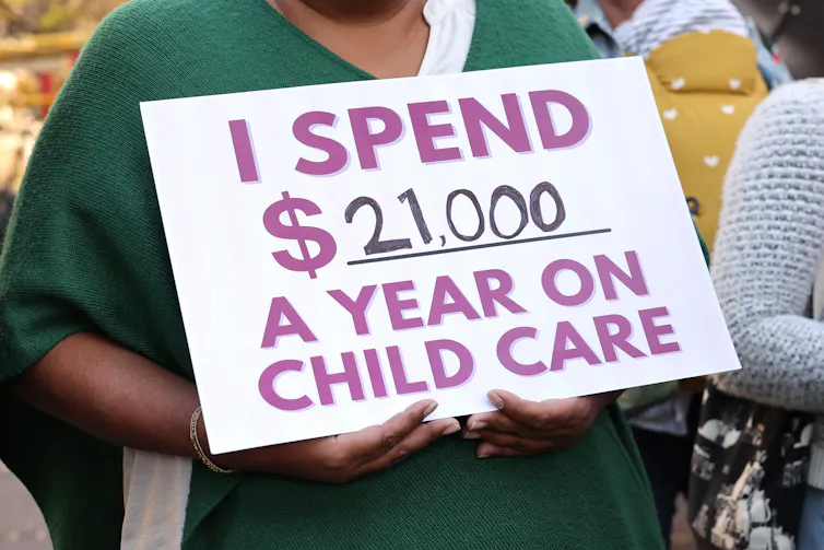 A person wearing a green shirt holds a sign that says in purple and black 'I spend $21,000 a year on child care.'